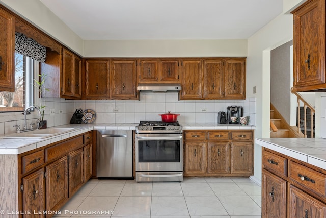 kitchen with under cabinet range hood, a sink, backsplash, tile countertops, and stainless steel appliances