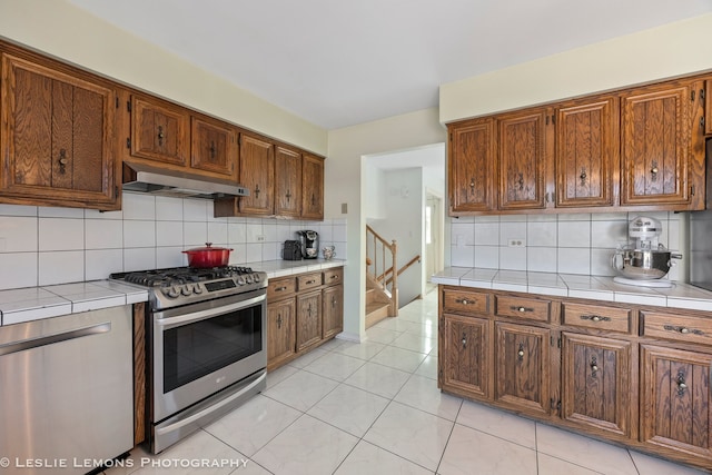 kitchen with tile countertops, light tile patterned floors, stainless steel appliances, under cabinet range hood, and tasteful backsplash