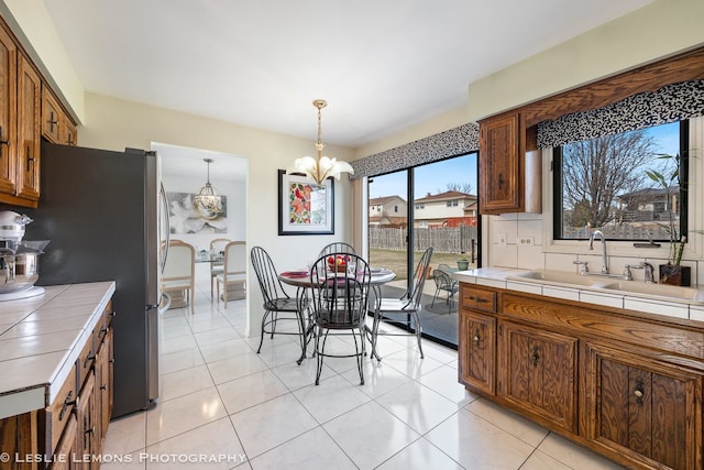 kitchen featuring tile countertops, freestanding refrigerator, a sink, hanging light fixtures, and a notable chandelier