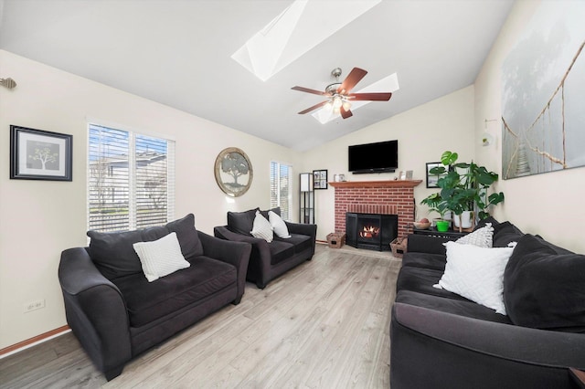 living area with baseboards, vaulted ceiling with skylight, light wood-style flooring, a ceiling fan, and a brick fireplace