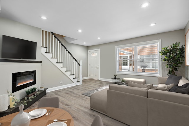 living area with stairs, a glass covered fireplace, wood finished floors, baseboards, and recessed lighting