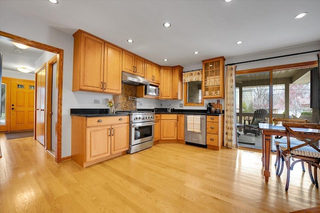 kitchen with glass insert cabinets, dark countertops, under cabinet range hood, light wood-style floors, and stainless steel appliances