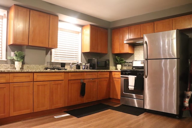 kitchen featuring light stone counters, brown cabinetry, stainless steel appliances, under cabinet range hood, and light wood-type flooring