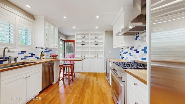 kitchen with wall chimney range hood, high end appliances, open shelves, a sink, and butcher block countertops