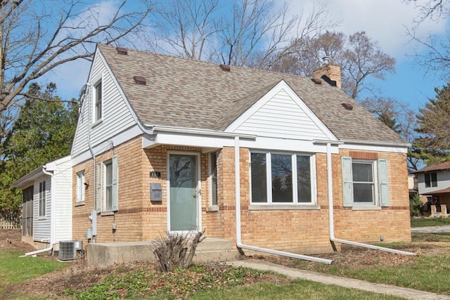 view of front facade featuring cooling unit, brick siding, a shingled roof, and a chimney