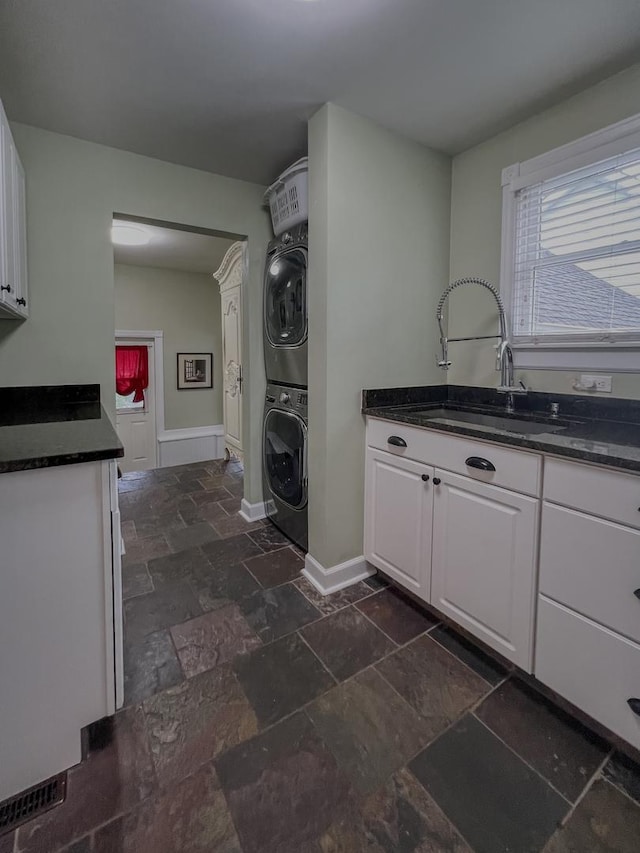 laundry area featuring laundry area, stone tile floors, stacked washer / dryer, baseboards, and a sink