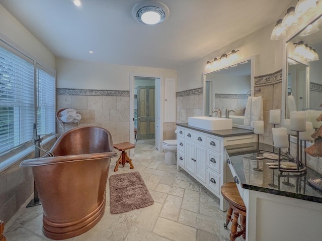 bathroom with tile walls, vanity, toilet, stone tile flooring, and a soaking tub