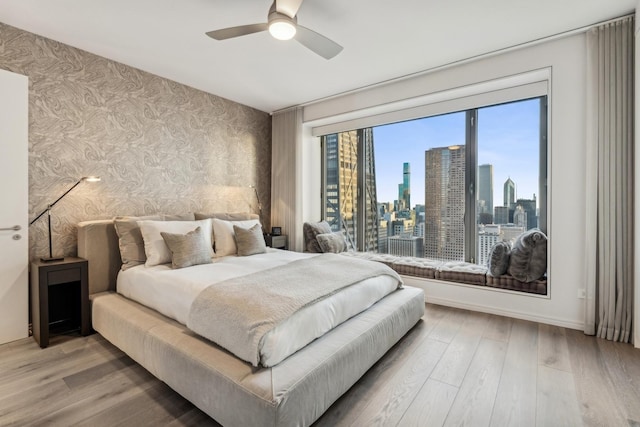 bedroom with baseboards, wallpapered walls, a city view, a ceiling fan, and wood finished floors