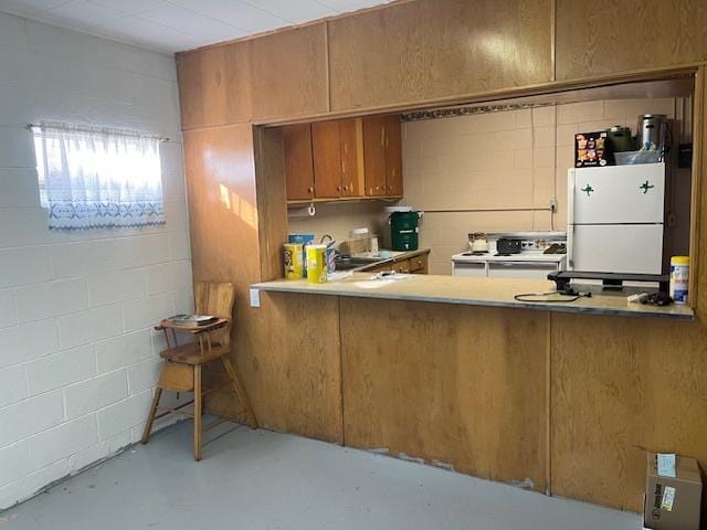 kitchen featuring concrete block wall, finished concrete floors, stove, brown cabinetry, and freestanding refrigerator