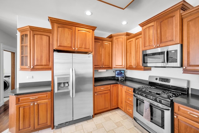 kitchen with appliances with stainless steel finishes, glass insert cabinets, washer / dryer, dark countertops, and recessed lighting