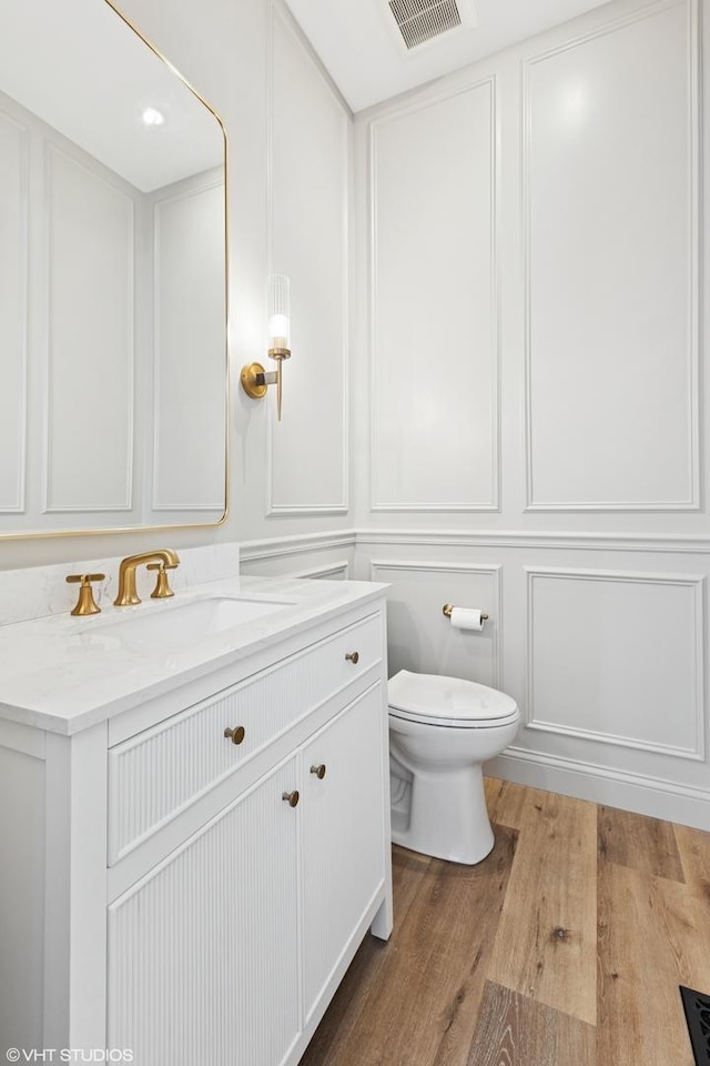 bathroom with toilet, visible vents, vanity, a decorative wall, and wood finished floors