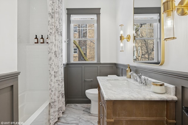 full bathroom featuring toilet, a decorative wall, wainscoting, marble finish floor, and vanity
