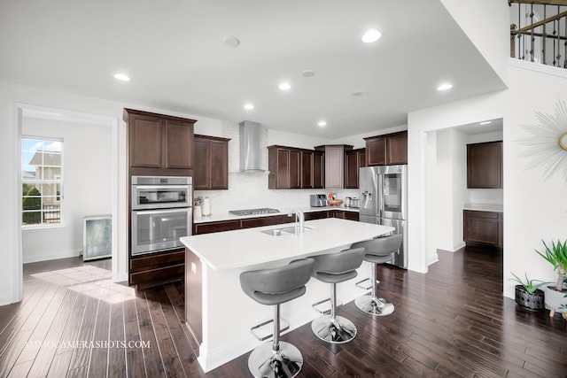 kitchen featuring wall chimney exhaust hood, a kitchen island with sink, dark wood-style flooring, a sink, and stainless steel appliances