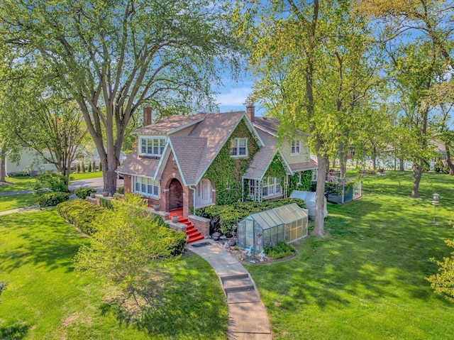 tudor-style house featuring a greenhouse, an outbuilding, a chimney, a front yard, and brick siding