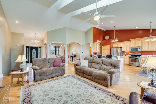 living room featuring high vaulted ceiling, light wood-type flooring, ceiling fan with notable chandelier, arched walkways, and ornate columns