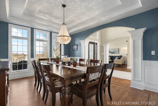 dining room featuring a chandelier, a raised ceiling, wood finished floors, arched walkways, and ornate columns
