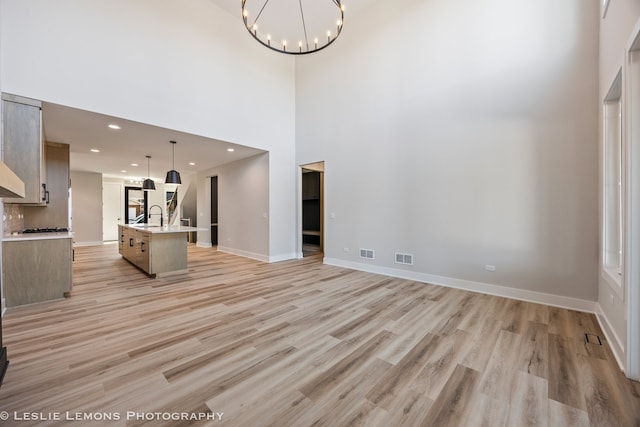 unfurnished living room featuring a towering ceiling, light wood finished floors, visible vents, baseboards, and a notable chandelier