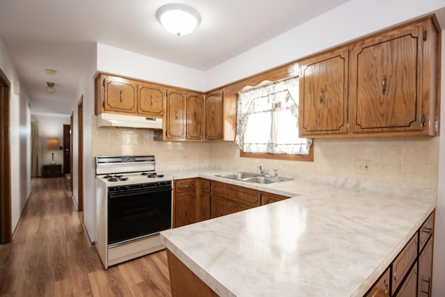 kitchen with light countertops, a sink, brown cabinets, under cabinet range hood, and white gas stove