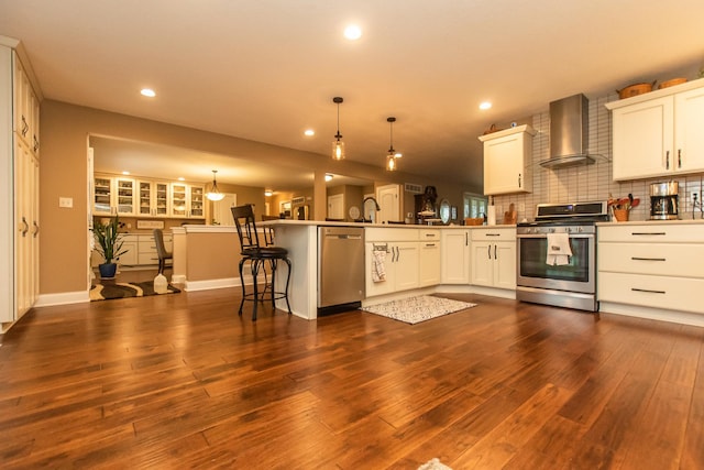 kitchen with dark wood-type flooring, backsplash, a peninsula, appliances with stainless steel finishes, and wall chimney exhaust hood