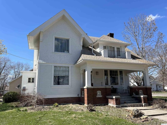 view of front of property featuring roof with shingles, a balcony, brick siding, a porch, and a chimney