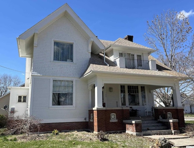 view of front of home with a balcony, a chimney, brick siding, a shingled roof, and a porch