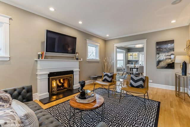 living area featuring light wood-style floors, crown molding, baseboards, recessed lighting, and a glass covered fireplace