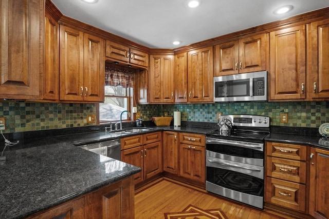 kitchen with brown cabinets, a sink, dark stone countertops, stainless steel appliances, and light wood-style flooring