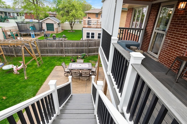 wooden deck featuring outdoor dining area, a trampoline, a patio area, a fenced backyard, and a yard