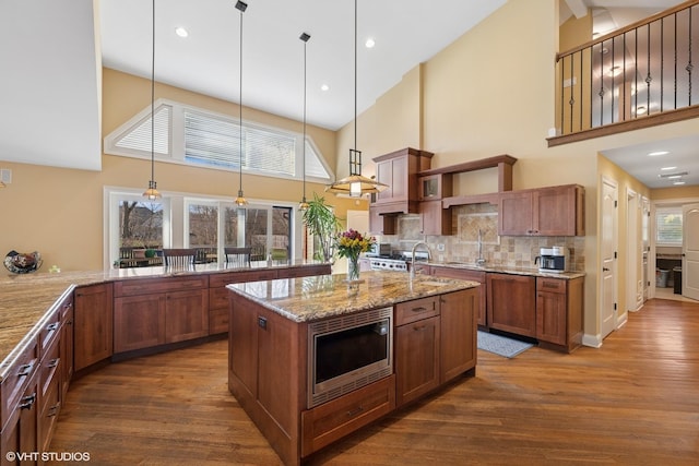 kitchen with high vaulted ceiling, light stone countertops, a kitchen island with sink, stainless steel microwave, and dark wood-style flooring