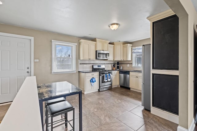 kitchen with light tile patterned floors, backsplash, cream cabinetry, dark countertops, and stainless steel appliances