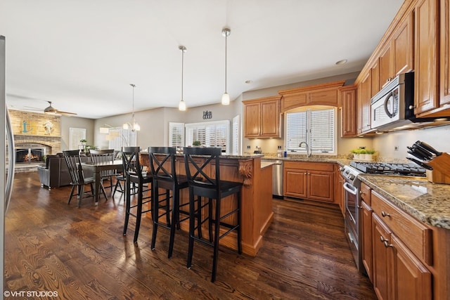 kitchen with ceiling fan with notable chandelier, a breakfast bar area, a fireplace, appliances with stainless steel finishes, and dark wood-style floors
