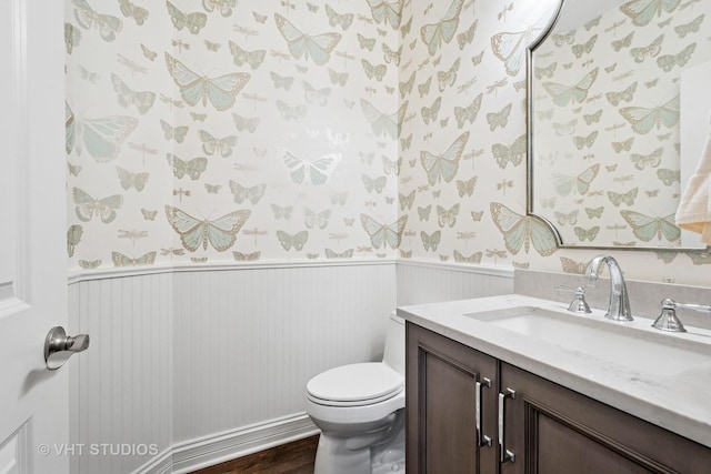 bathroom featuring toilet, wallpapered walls, vanity, and wainscoting