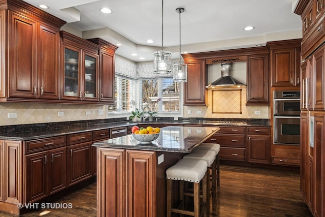 kitchen with appliances with stainless steel finishes, dark wood-style flooring, wall chimney range hood, and a kitchen island