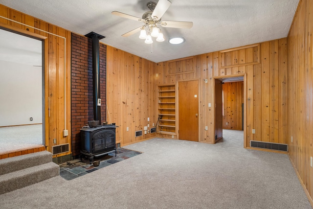 unfurnished living room with wooden walls, visible vents, carpet flooring, and a wood stove