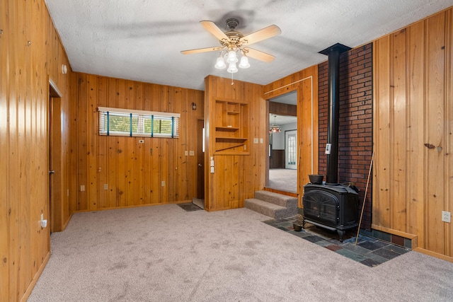 unfurnished living room featuring wood walls, ceiling fan, carpet flooring, a wood stove, and a textured ceiling