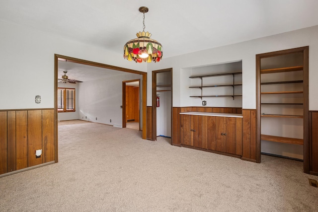 unfurnished dining area featuring a wainscoted wall, wooden walls, light carpet, a ceiling fan, and built in shelves