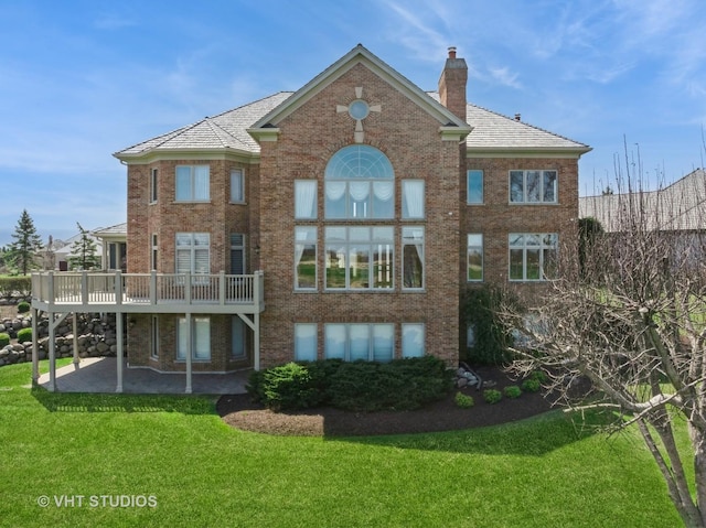 back of property with a chimney, brick siding, a wooden deck, a lawn, and a patio
