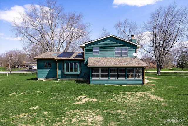 back of property with a chimney, a shingled roof, a yard, solar panels, and a sunroom