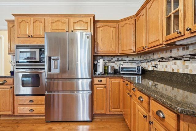 kitchen with crown molding, backsplash, glass insert cabinets, dark stone countertops, and stainless steel appliances