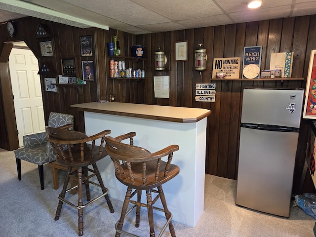bar with wood walls, a bar, light colored carpet, and freestanding refrigerator