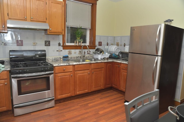 kitchen featuring a sink, light stone countertops, appliances with stainless steel finishes, under cabinet range hood, and dark wood-style flooring