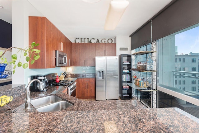kitchen with appliances with stainless steel finishes, tasteful backsplash, brown cabinets, a sink, and dark stone countertops