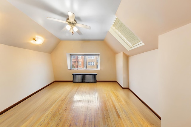 additional living space featuring baseboards, radiator, light wood finished floors, lofted ceiling with skylight, and a ceiling fan
