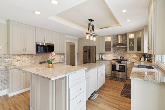kitchen with a tray ceiling, light wood-style flooring, wall chimney range hood, a kitchen island, and appliances with stainless steel finishes