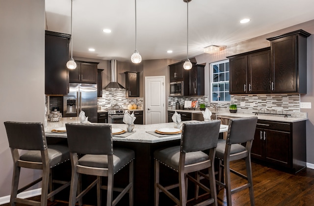 kitchen featuring wall chimney range hood, a kitchen breakfast bar, dark brown cabinetry, stainless steel appliances, and dark wood-type flooring