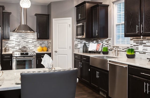 kitchen with light stone counters, wall chimney range hood, a healthy amount of sunlight, stainless steel appliances, and a sink