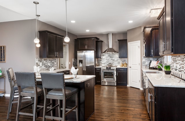 kitchen featuring wall chimney exhaust hood, dark brown cabinetry, dark wood-type flooring, a peninsula, and appliances with stainless steel finishes