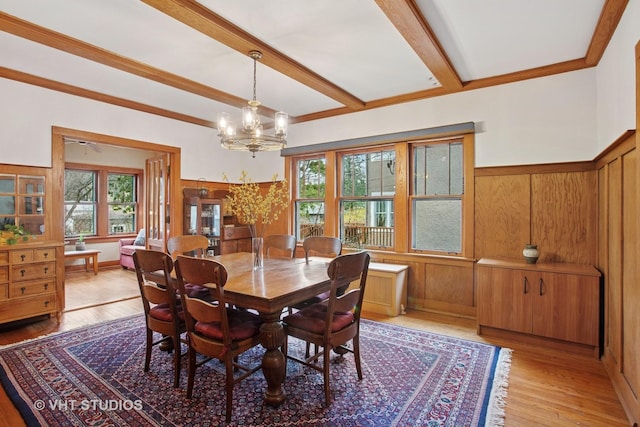 dining space with light wood-style floors, beamed ceiling, an inviting chandelier, wood walls, and wainscoting