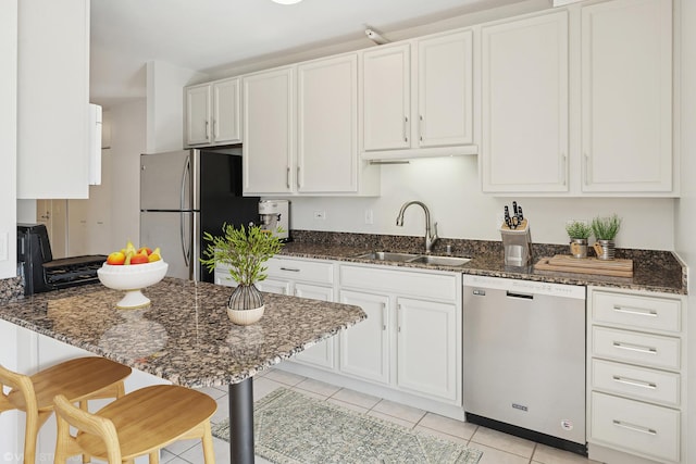 kitchen featuring stainless steel appliances, a sink, light tile patterned floors, dark stone counters, and white cabinetry