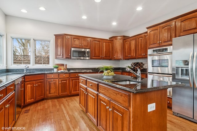 kitchen with light wood finished floors, a kitchen island with sink, stainless steel appliances, dark stone counters, and a sink
